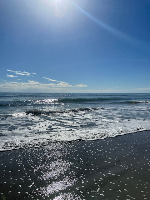 The sun is shining on the water as gentle waves crest quietly and roll onto the shore. The sky is blue with white clouds dotted here and there and also on the horizon.