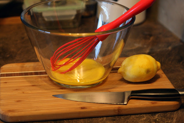 Egg yolk in a glass bowl, with a lemon and a knife on a bamboo cutting board. 