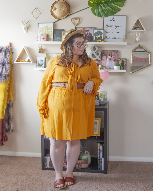 An outfit consisting of a straw boater hat, a yellow long sleeve bell sleeve with ties mini wrap dress with a wide brown braided belt and brown slide sandals.