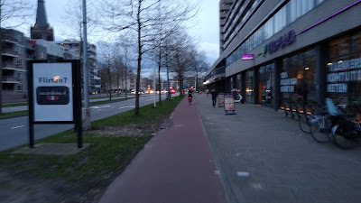 A wide street with a red one-way cycle track with a grass verge left and a road beyond and a footway to the right in front of shops. It's dusk.