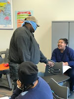 A blindfolded male student reaches to touch a chair in front of a female student who is seated