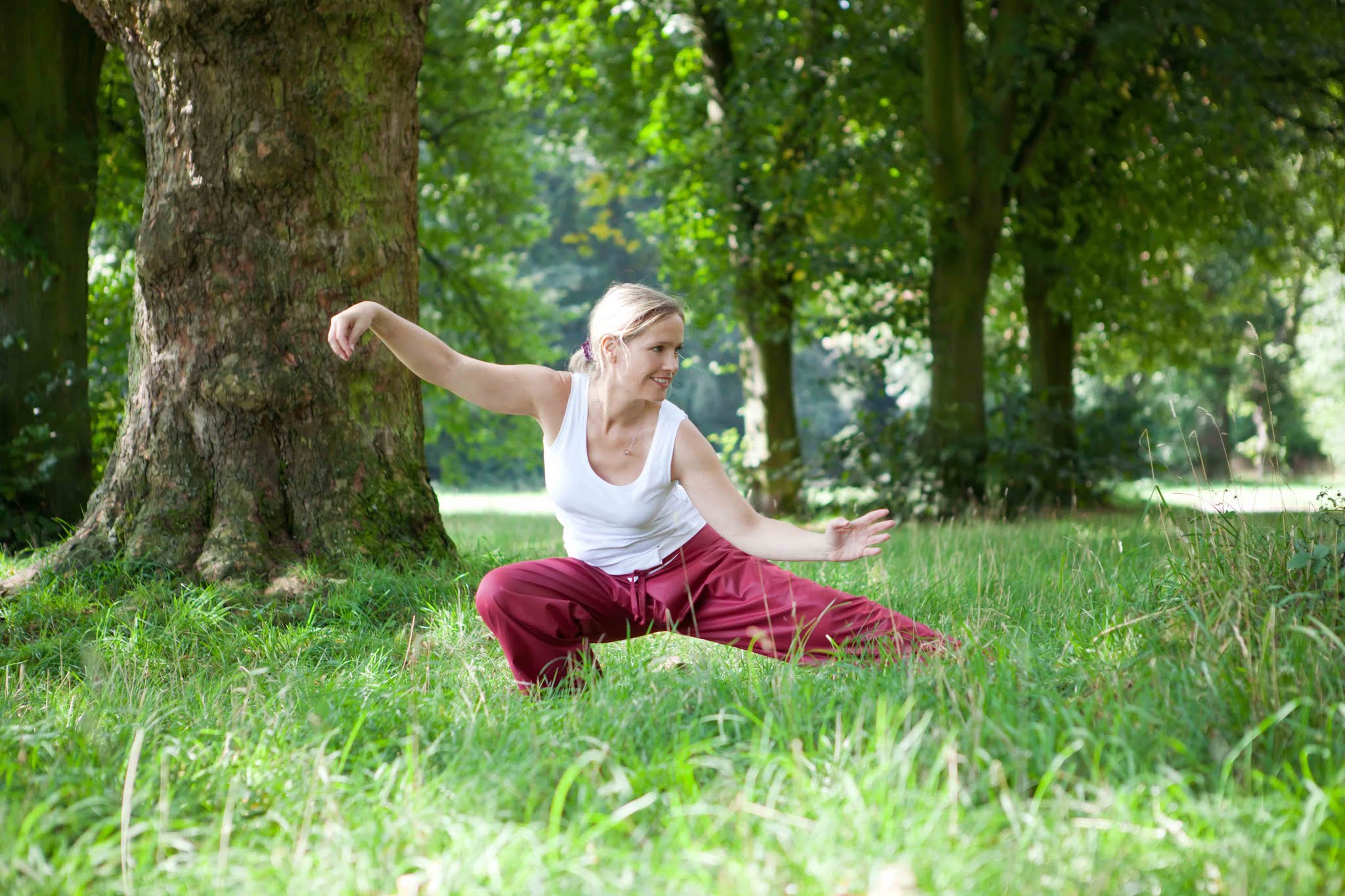 Frau in roter Hose und weißem Shirt bei  Tai Chi auf grünem Gras unter Bäumen