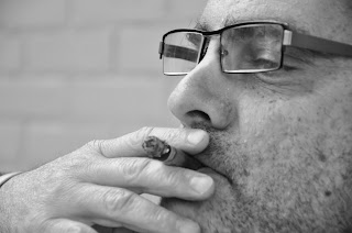 Homem com óculos e barba fumando um cigarro.