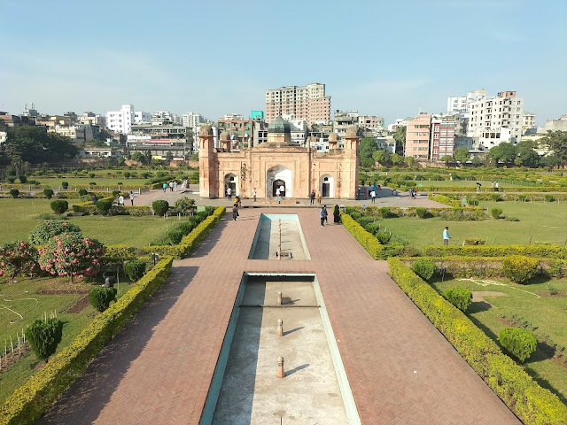 Path with empty water fountains in centre and gardens on either side leading to domed structure with reddish walls and apartment buildings visible behind