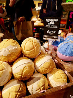 A selection of a mix of orange and yellow spherical bath bombs with patterns all over on a large wide rectangular light brown shelf with a black rectangular board saying Turmeric Latte on a bright background