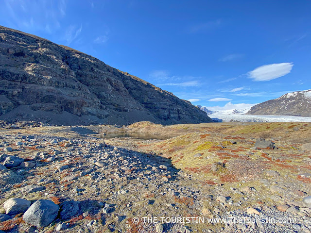 A bright blue sky with a few white clouds over mountainous terrain in front of a large body of glacial ice extending across and between a mountain range.