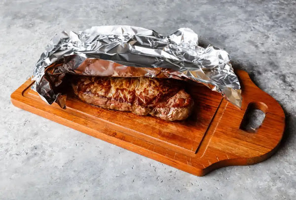 Grilled steak resting on a wooden cutting board, lightly tented with aluminum foil to retain juices.