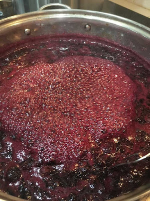 A silver saucepan full of deep-magenta jam at a rolling boil, with a thick mass of bubbles in the center.