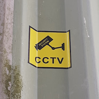 Bright yellow sticker on a corrugated steel wall: reads '24 hour CCTV'