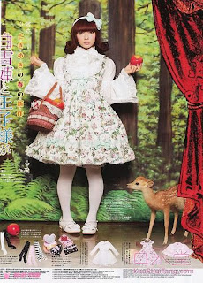 Misako Aoki modeling the Blooming Snow White jumperskirt in green colourway. She is wearing a blouse with princess sleeves, tights and flat shoes. She is holding a basket of apples. There is a fake deer next to her. She is standing in front of a forest background. There is a red curtain off to the side.