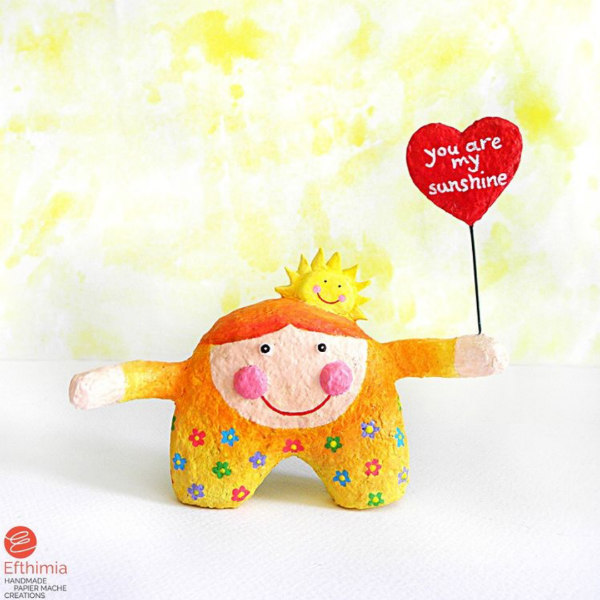 whimsical paper mache girl figurine with outstretched arms holding a You Are My Sunshine heart-shaped sign
