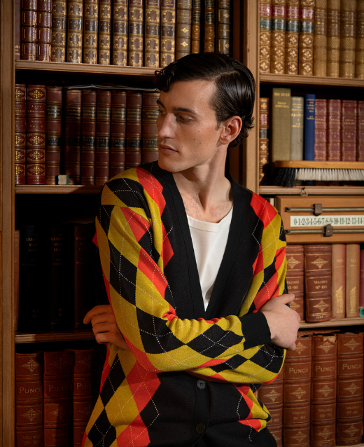 Male Model leaning against an old style bookcase wearing designer clothing. The clothing consists of Stella McCartney printed cardigan and white low neck top