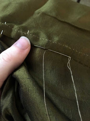 A close-up of a seam in green-gold silk being flat-felled, with the tan-threaded needle inserted for the next slip stitch.