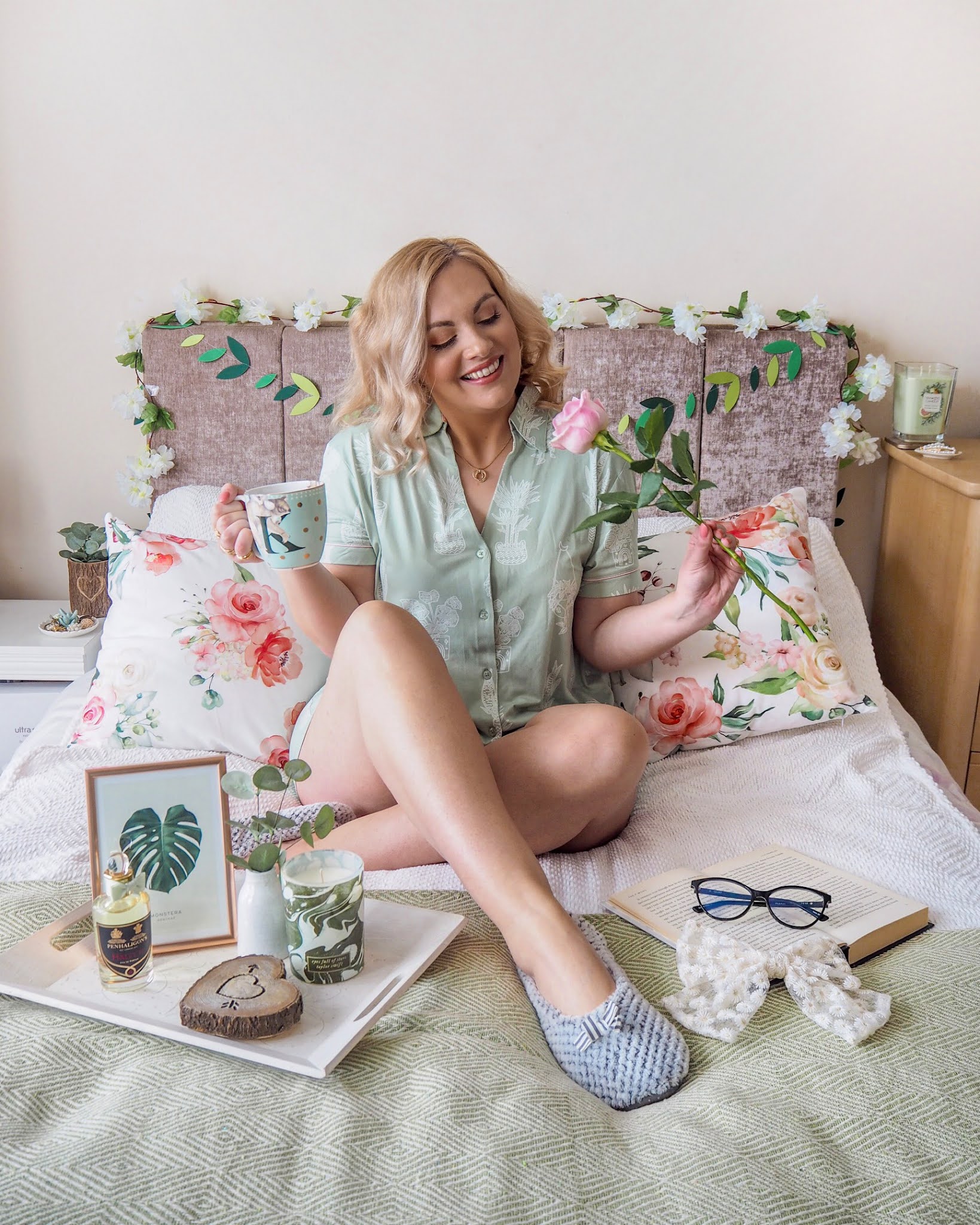 Spring Photography Props & Home Decor, Girl sitting on a bed in sage green pyjamas holding a rose and a mug of tea there are lots of floral spring props like cushions and flower garlands around her.