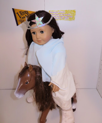 An American Girl doll in a white dress with blue cape sitting astride a horse