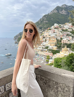 A person in sunglasses smiles, standing by a stone wall overlooking a coastal town with colorful buildings and boats on the water.
