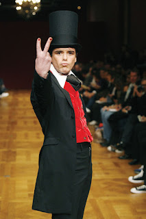 Sebastian Horsley on a catwalk wearing a black suit and top hat with a red waistcoat while making a traditionally offensive 'V-sign' with his fingers to the camera