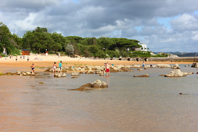 naznaczane wystającymi kamieniemi płytkie wody zatoki przy niewielkiej plaży z parkiem na półwysepie La Magdalena w tle 