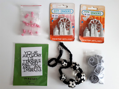 Flat lay of a fridge magnet, a necklace with large daisy beads on it, 2 sets of erasers in the shape of city buidlings, some clay cutters and a bag of one-twelfth scale baby bottles.