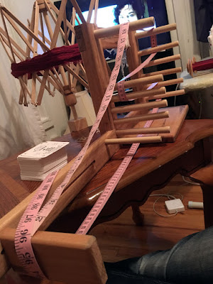 A wooden tablet-weaving loom with a pink measuring tape wrapped around a number of the pegs, with a stack of white tablet-weaving cards to the left of the loom on the table, and a wooden swift holding a thick hank of extremely fine deep garnet thread.