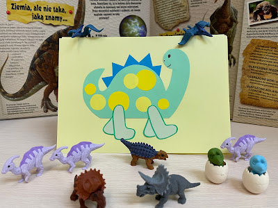 Na pierwszym planie kilka plastikowych figurek dinozaurów. Za nimi, oparta o książkę o dinozaurach, znajduje się praca plastyczna - na jasnozielonej kartce przyklejony zielony dinozaur z niebieskimi kolcami.