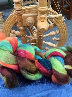 A thick braid of spinning fiber in mixed shades of magenta, orange, green, teal, and brown, on the photographer's lap in front of a small upright wooden spinning wheel.