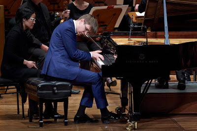 Photo of a man in a blue suit playing a grand piano with a woman in black wearing white glasses seated to his left.