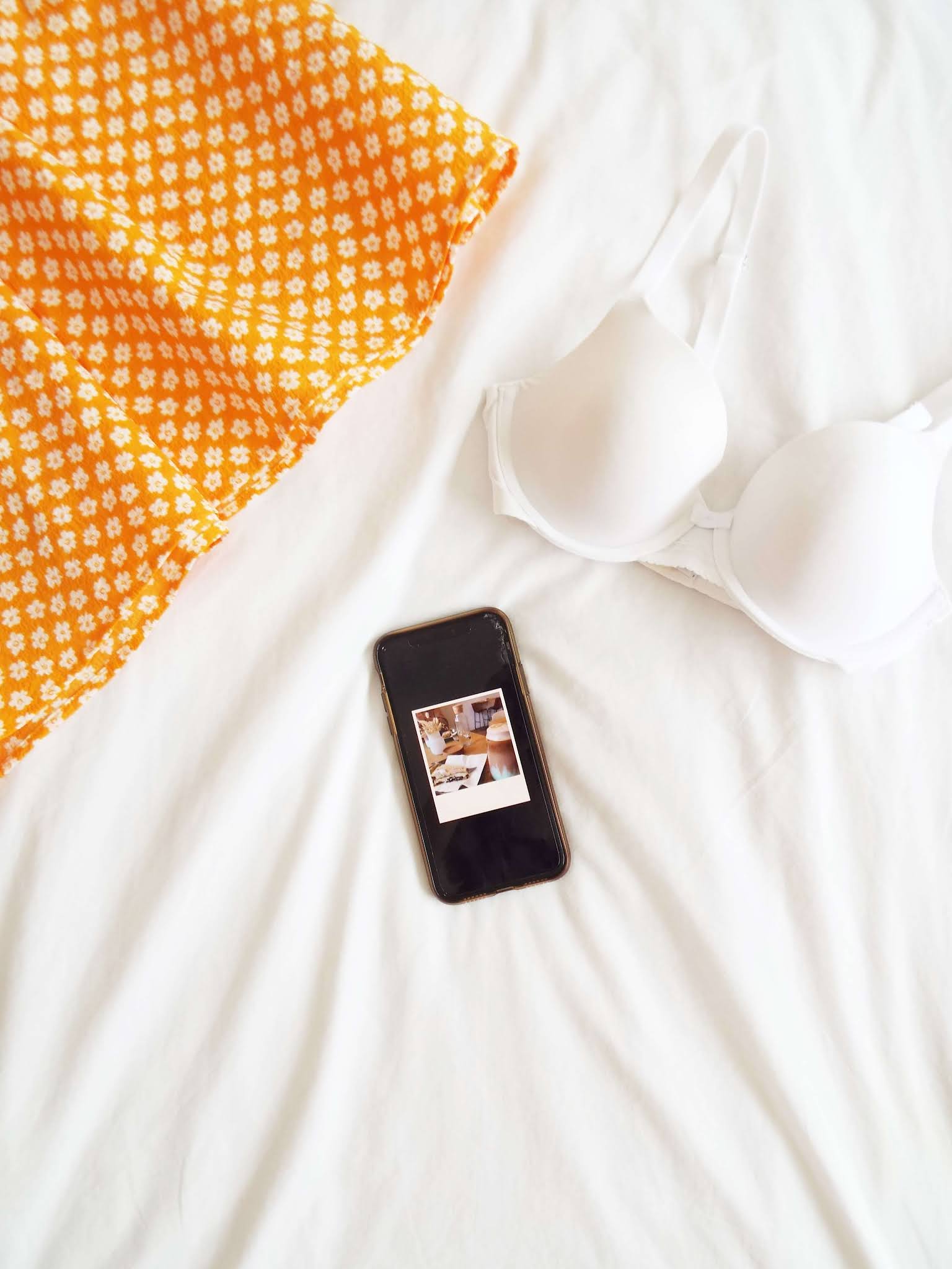 Orange floral dress, white bra and phone with little polaroid picture displayed on it, with cookie pie and an iced latte, lay on white bedding.