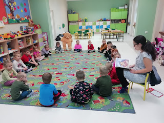 Sala przedszkolna. Dzieci siedzą na dywanie. W tym samym okręgu siedzi bibliotekarka oraz pluszowy miś