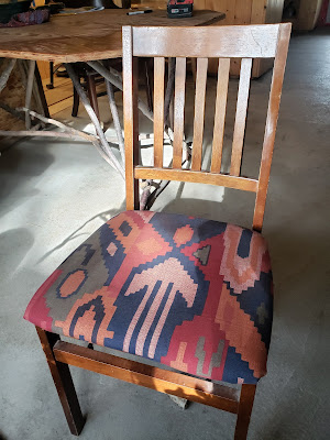 Image shows a wooden chair with a newly upholstered seat cushion on it. The fabric on the cushion is maybe an Aztec-inspired geometric design with dulled/neutral colours - burgundy, mossy green, dark navy blue, a rusty orange, and a salmony pink colour. The rest of the chair is a standard wood chair with wooden columns for a backrest.