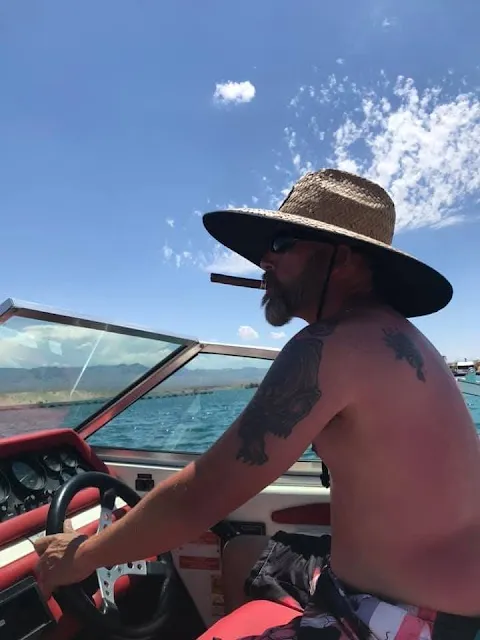 Side view of a man driving a boat with a straw hat on shirtless tattoos on him and swim trunks smoking a cigar