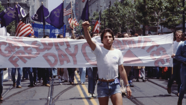 A film still of Gay USA. A man stands in front of a protest with his fist in the air. He's standing in front of a banner that reads Gay Freedom Day Committee.