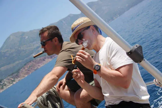 1/2 two men driving a boat and smoking cigars with beautiful mountains and blue water in thy background