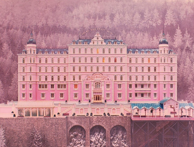 Cena de "O Grande Hotel Budapeste". Mostra uma luxuosa fachada de hotel. A parede é cor-de-rosa e o telhado azul.