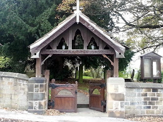 A wooden lychgate over a double wooden gate set in a stone wall.  Behind are trees in a graveyard.