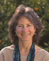 Head-shot of a light-skinned middle-aged thin woman with wavy brown hair and rosy cheeks, smiling broadly. Her eyes look like they're looking right at you. She's wearing pearl drop earrings, a blue-and-purple scarf, and a tan sweater.