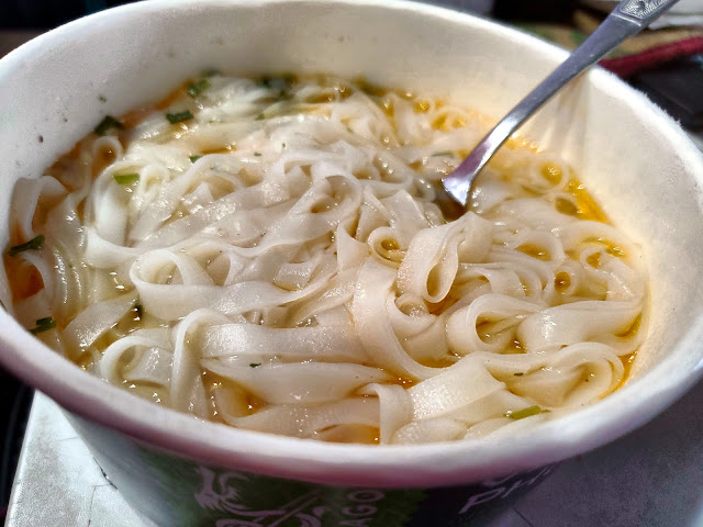 A cardboard bowl with rice noodles, soup and a fork.