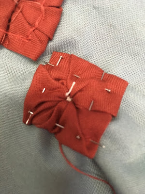 A small, roughly folded square of red fabric, pinned along the edges of the square to grey-blue fabric, with messy pleats running to the center of the square.