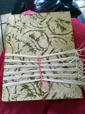 A small white and gold notebook sitting on a pink-skirted lap, with six bands of white cord tied around it. All the knots are at the right side, and the bands are concentrated at the bottom of the notebook, separated by equal amounts of empty space. A pink cord cinches them together slightly at the center, with a knot at the bottom.