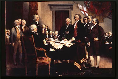 American founding fathers, John Adams, Thomas Jefferson, and others standing at a table on which is the Declaration of Independence