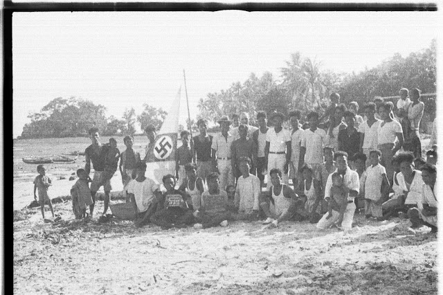 Group of people posing for a photo on a beach, with a canoe with a large swastika on its sail.