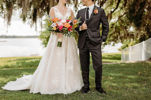 Bride in white lace wedding dress holding colorful bouquet with groom in black suit with colorful boutonniere