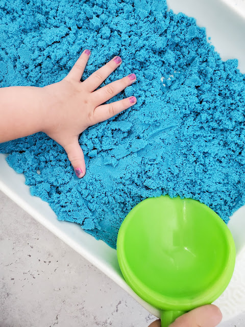 a child's hand playing with the sand in a white casserole dish.