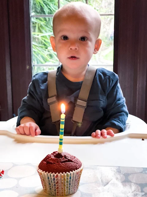 Baby Boy sitting in a high chair with a red velvet cupcake in front of him. The cake has a single lit candle in 