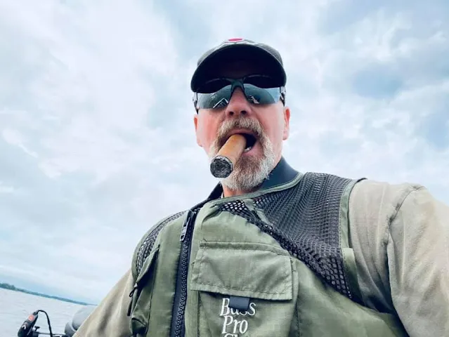 Army dude wearing sunglasses and smoking a cigar on a boat smiling with a Gray beard