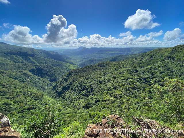 Lush green vegetation of a tropical island rainforest and in various locations pointy mountains covered in vegetation, under a soft blue sky with a few white fluffy cumulus clouds.