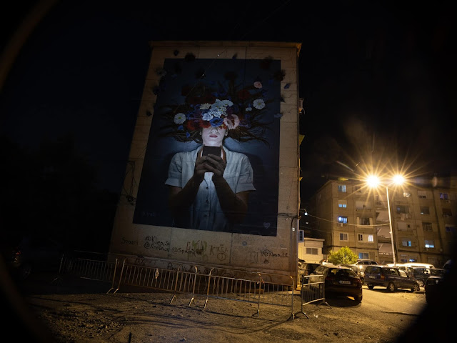 The Fabian Bane's mural in Tirana at night where a girl is painted looking at her phone and the upper half of her face is covered with colorful flowers
