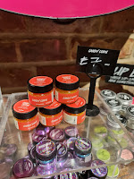 A photo of a light brown rectangular shelf filled with a stack of cylindrical glass jars filled with orange scrub  with black circular lids with a orange circular label that says candy corn lip scrub in white font on a bright background