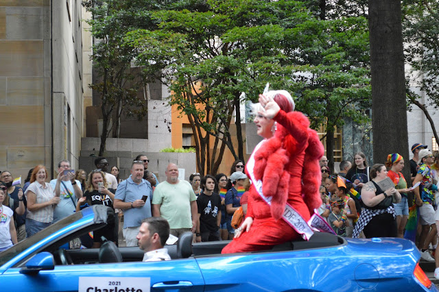 Drag queen in all red sitting on the back of a convertable waving to the crowd.