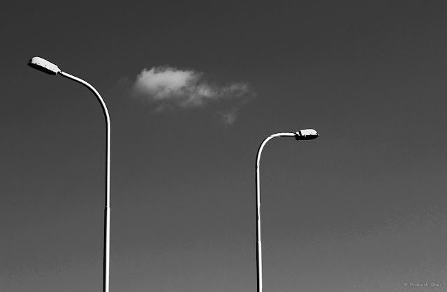 A black and white Minimalist Photo of two street lamps curved in the opposite direction split by a Minimalist White Cloud